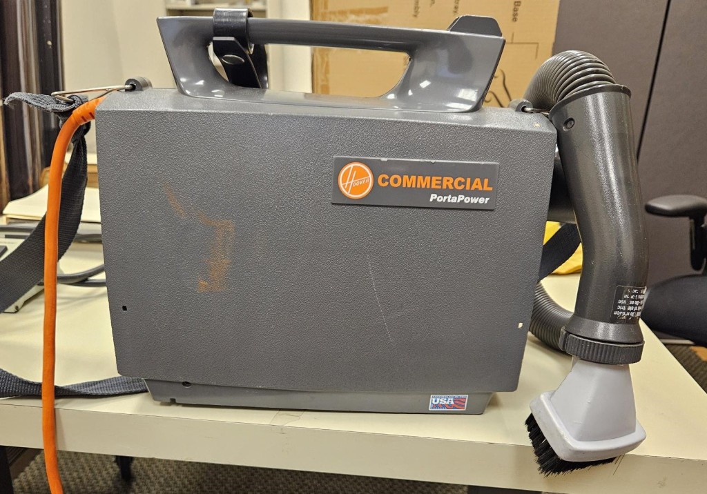A photograph of a commercial vacuum sitting on a worktable. The vacuum is a gray box with the words "Hoover Commercial PortaPower" written in orange and white on the side.
